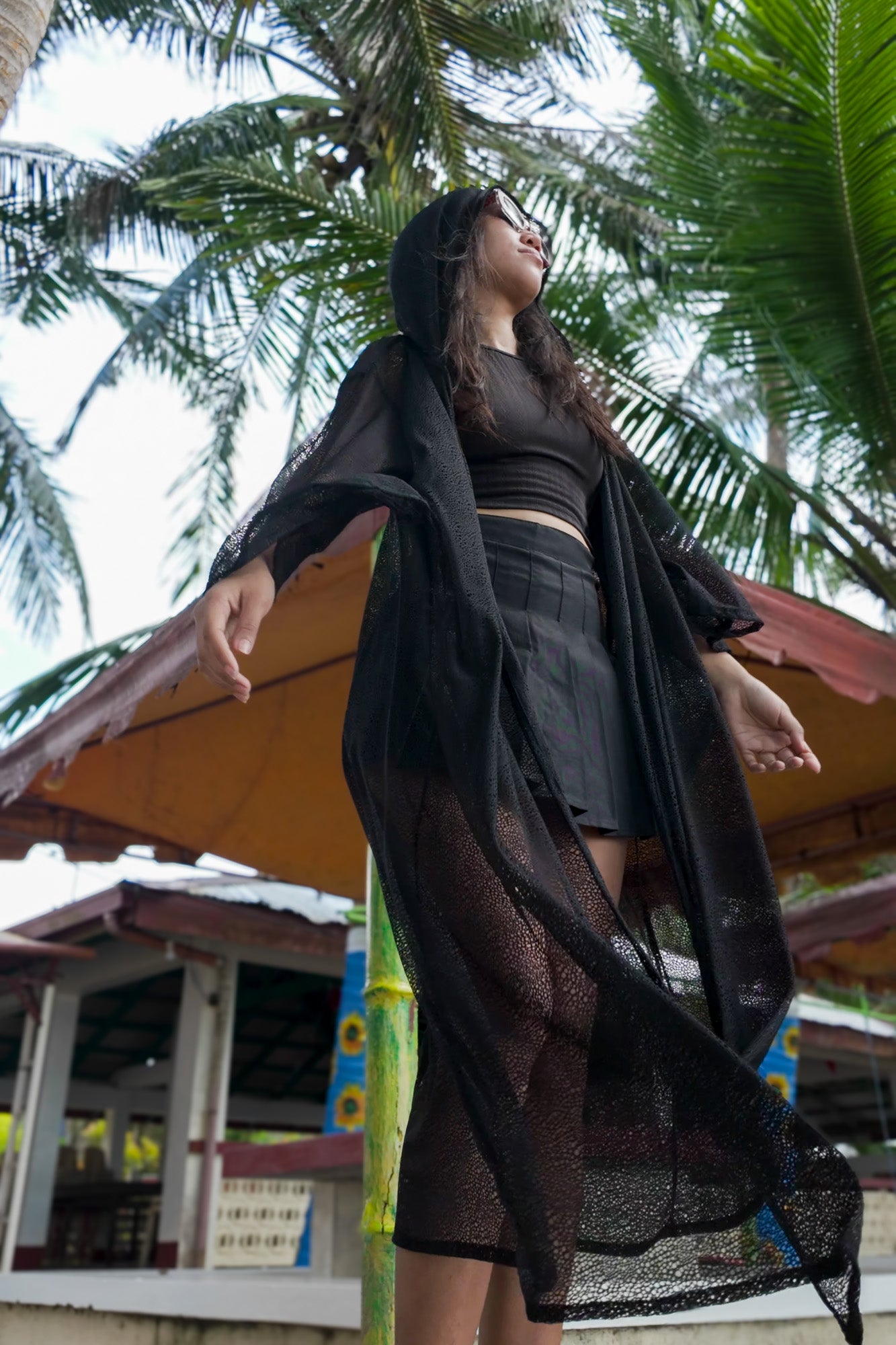 Woman in black outfit standing under a palm tree with a traditional hut in the background